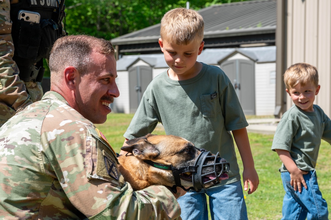Photo shows a military working dog handler holding a muzzled military working dog as a child pets the dog. There is another child smiling as he walks away from the dog.