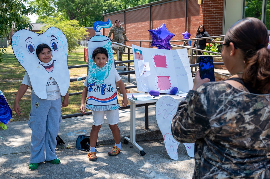 Photo shows the back of a mom holding up a phone to take a photo of her two children in front of her. The child on the left is wearing a tooth cardboard cutout and the child on the right is wearing a toothpaste cardboard cutout.
