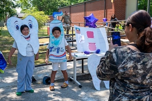 Photo shows the back of a mom holding up a phone to take a photo of her two children in front of her. The child on the left is wearing a tooth cardboard cutout and the child on the right is wearing a toothpaste cardboard cutout.