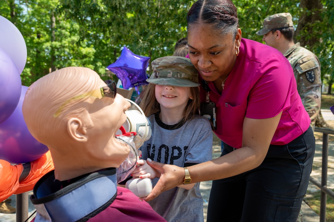 Photo shows a dental assistant holding a dental tool with a child pointing the tool towards a mannequin. The child is wearing a military hat.
