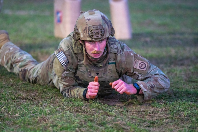 A man wearing a camouflage military uniform and helmet crawls through grass.