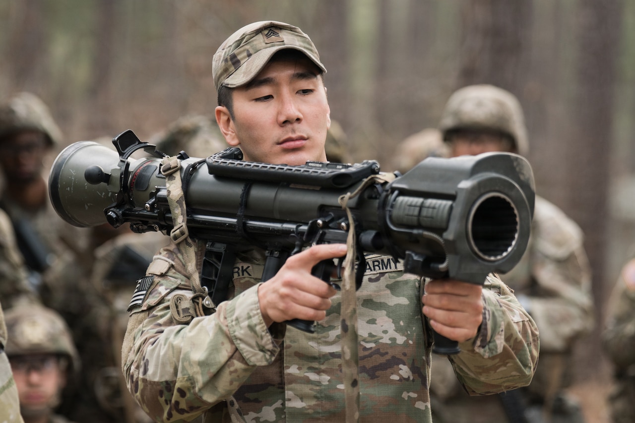 A man wearing a camouflage military uniform balances a weapon on his shoulder and prepares to shoot.