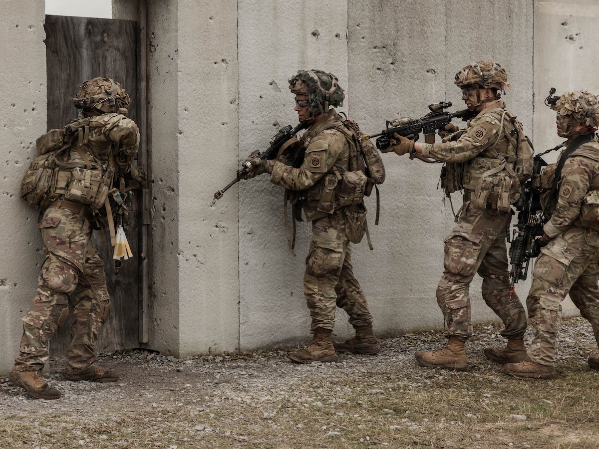 Three people wearing camouflage military uniforms and helmets hold rifles outside of a concrete structure as a person in similar attire looks inside the door.