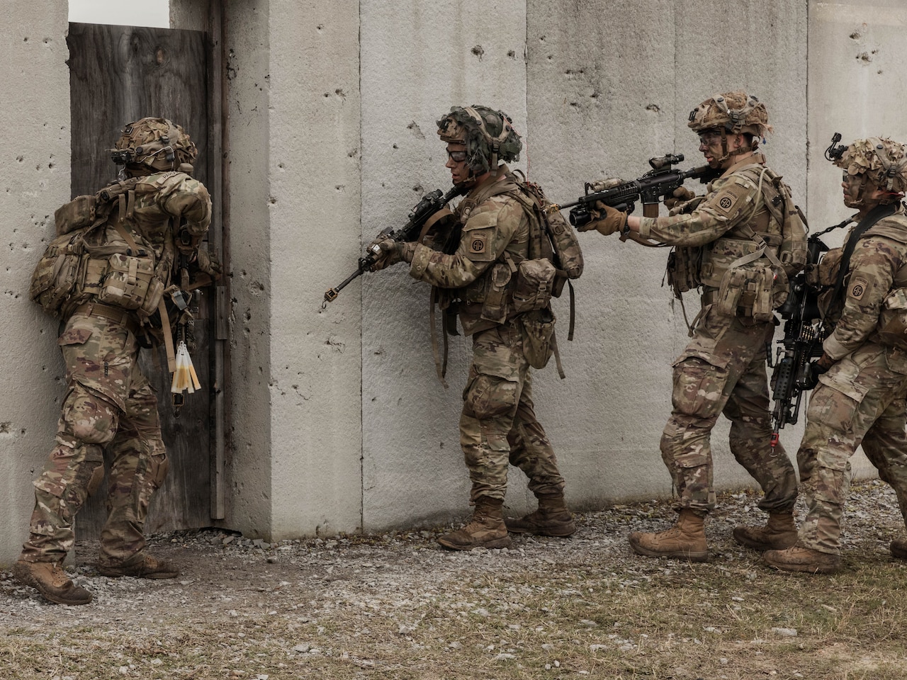 Three people wearing camouflage military uniforms and helmets hold rifles outside of a concrete structure as a person in similar attire looks inside the door.