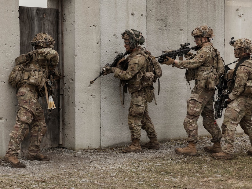 Three people wearing camouflage military uniforms and helmets hold rifles outside of a concrete structure as a person in similar attire looks inside the door.