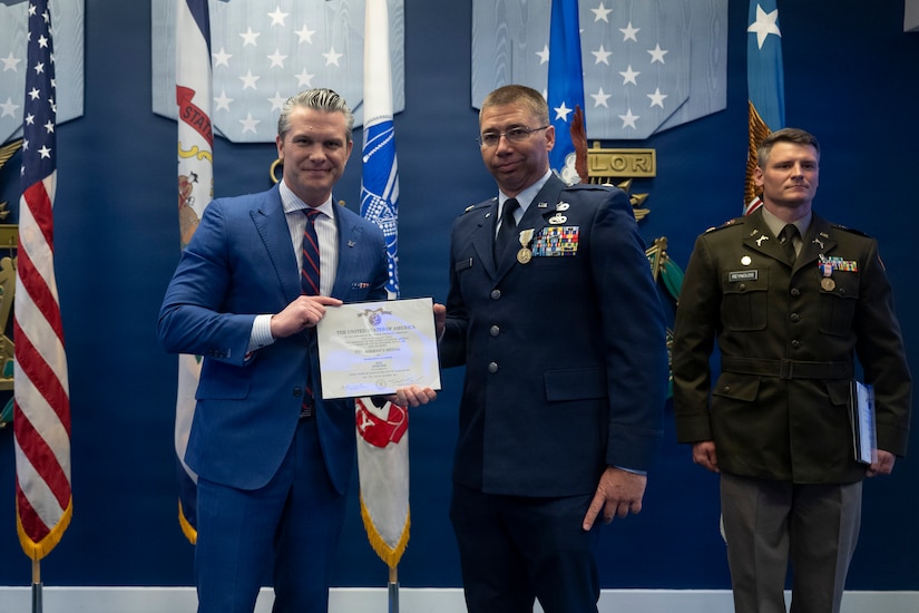 The secretary of war holds a certificate while posing in a photo with a service member as a fellow service member stands at attention to the right.