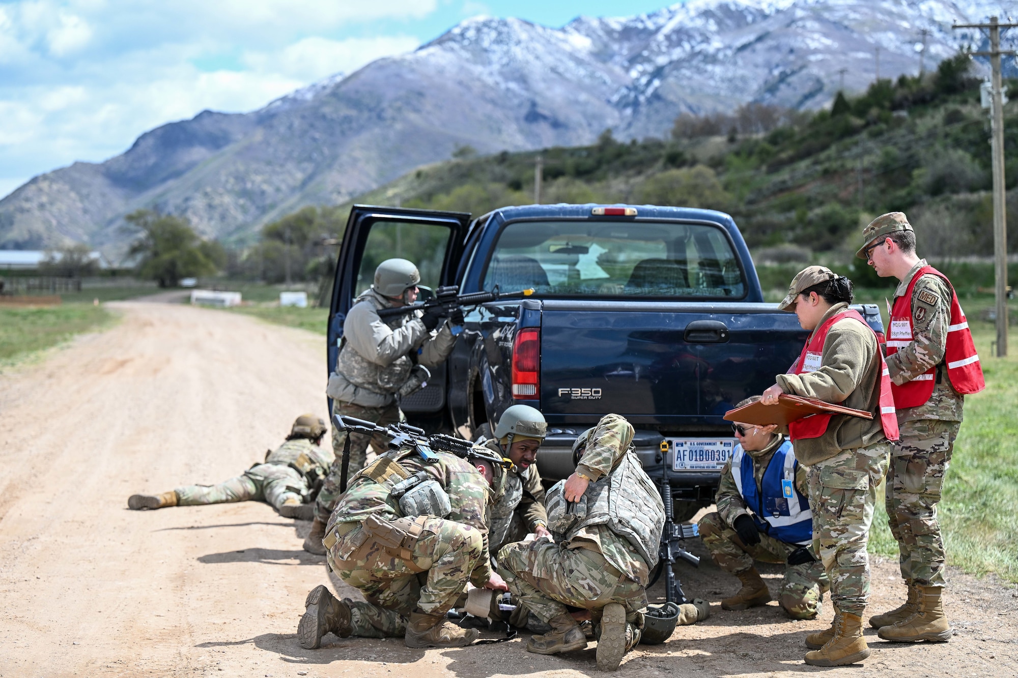 Airmen in camouflage gear take cover behind a blue pick up truck on a dirt road with mountain background during an exercise
