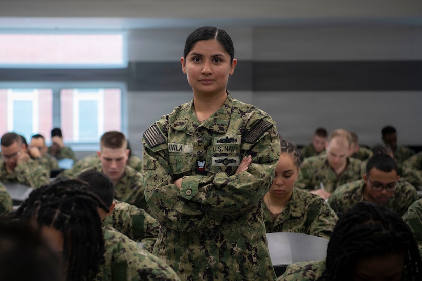 A sailor in a camouflage uniform stands with her arms crossed in a classroom setting, as sailors in similar attire sit at desks around her.