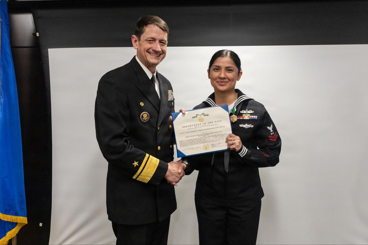 A person in a military dress uniform shakes the hand of a sailor as both of them hold a certificate and smile indoors.
