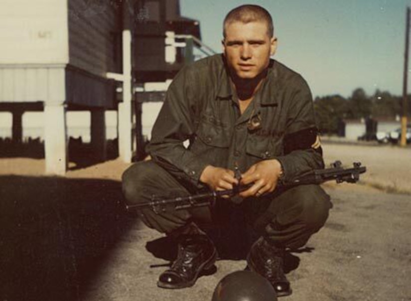 A man in a military uniform holds a weapon while squatting on the ground outside.
