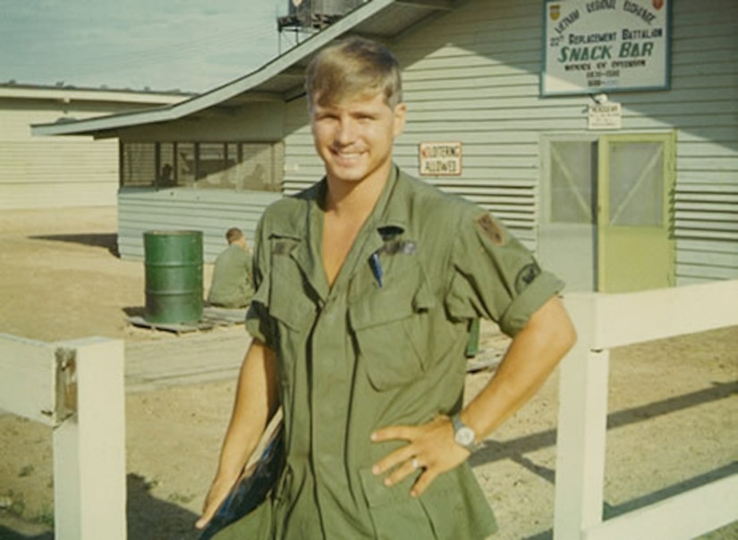 A man wearing a green military uniform smiles outside in front of a building with a sign over the door that reads Snack Bar.
