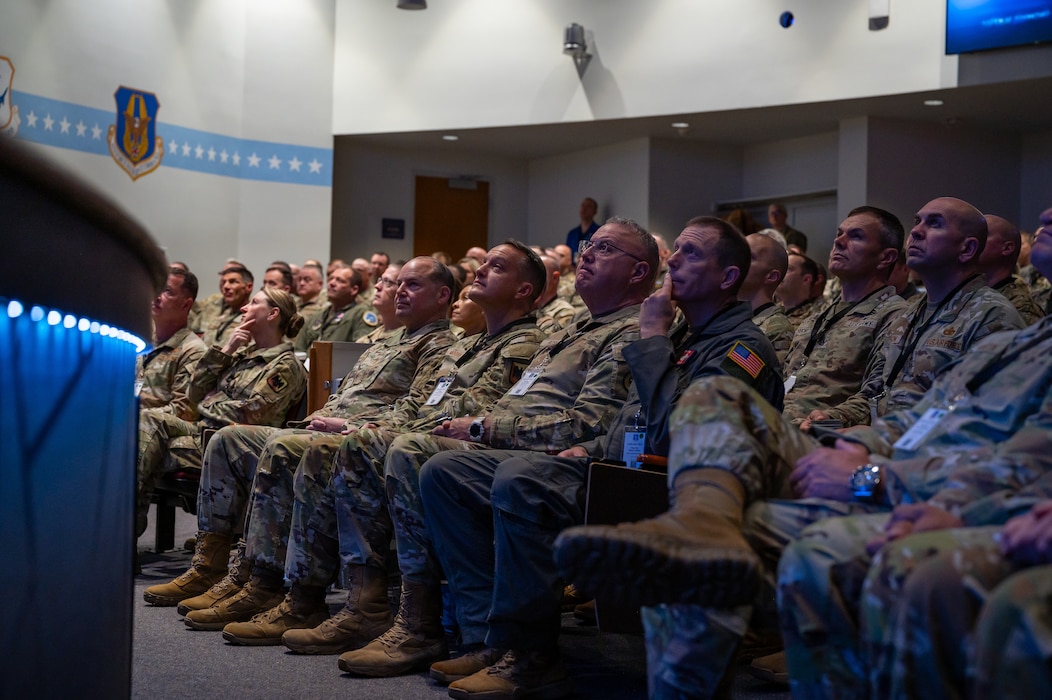 U.S. Airmen in OCP uniforms and flight suits sit in an audience in a dimly-lit auditorium during a moment of silence.