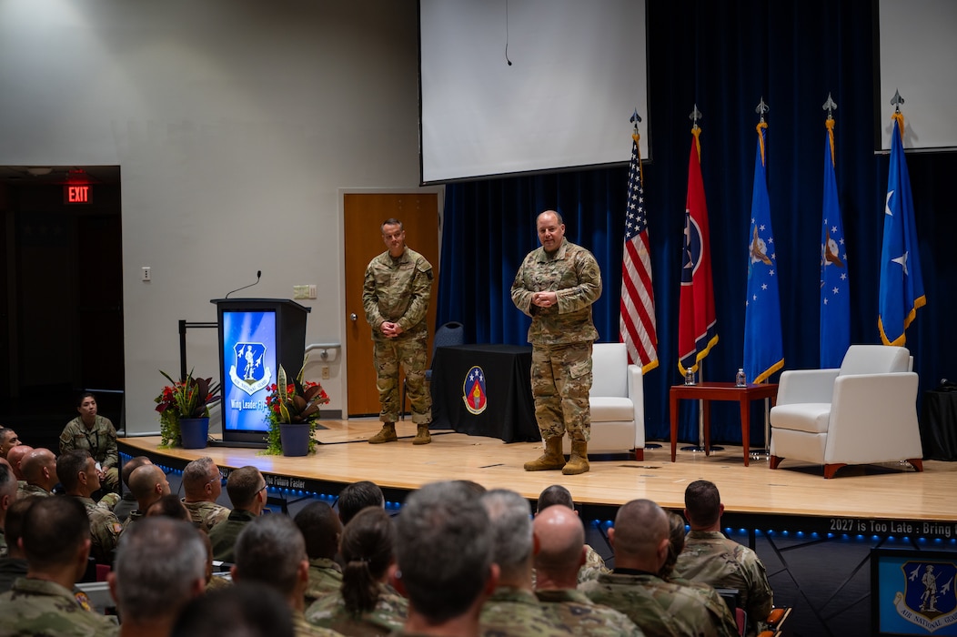 Two U.S. Airmen in OCP uniforms speak from a stage to an audience of U.S. Airmen in OCP uniforms and flight suits.