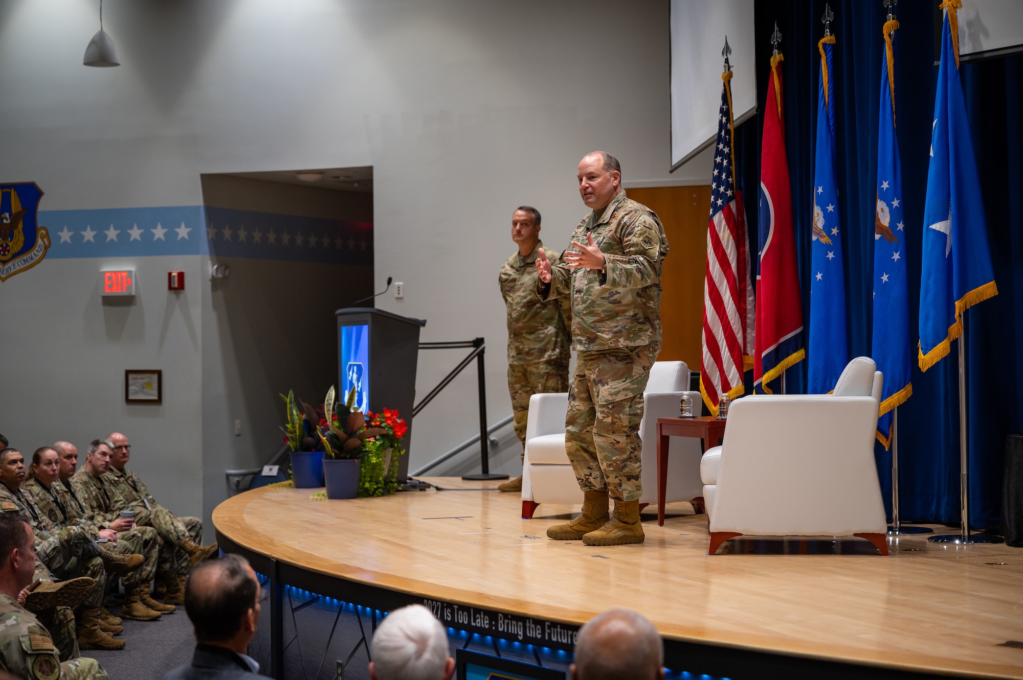 Two U.S. Airmen in OCP uniforms speak from a stage to an audience of U.S. Airmen in OCP uniforms and flight suits.