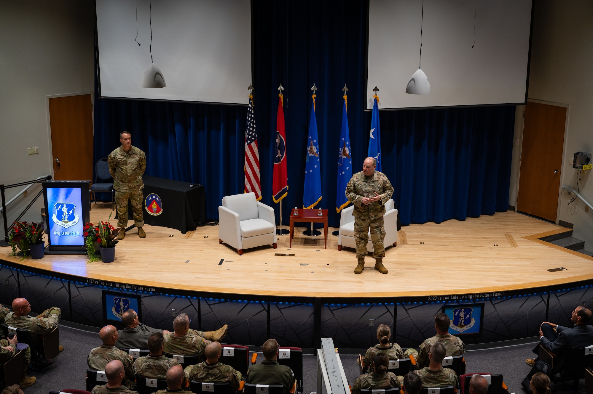 Two U.S. Airmen in OCP uniforms speak from a stage to an audience of U.S. Airmen in OCP uniforms and flight suits.