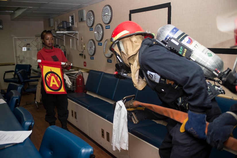 A sailor in firefighting gear holds a large hose and crouches in front of another sailor holding a red and yellow flag with an "A" on it indoors.