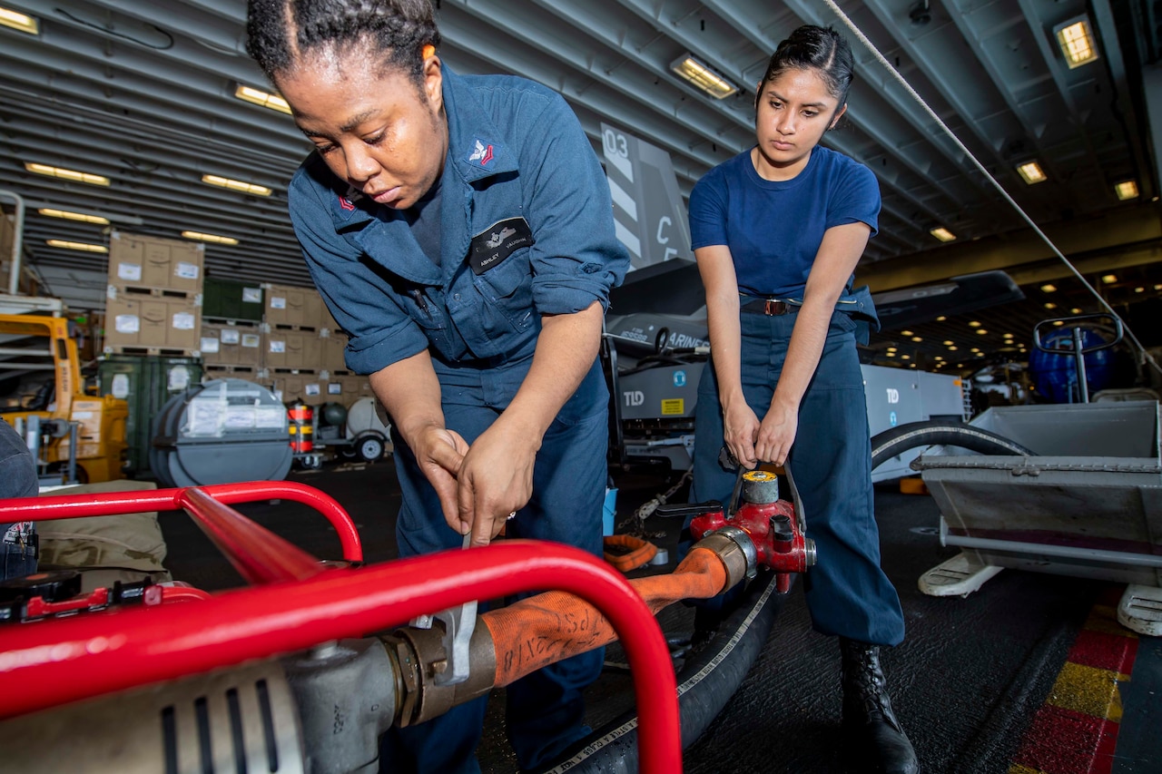 Two sailors, one using a wrench-like device to attach a hose to a pump as the other holds an attached piece of equipment, work in the hangar bay of a vessel.
