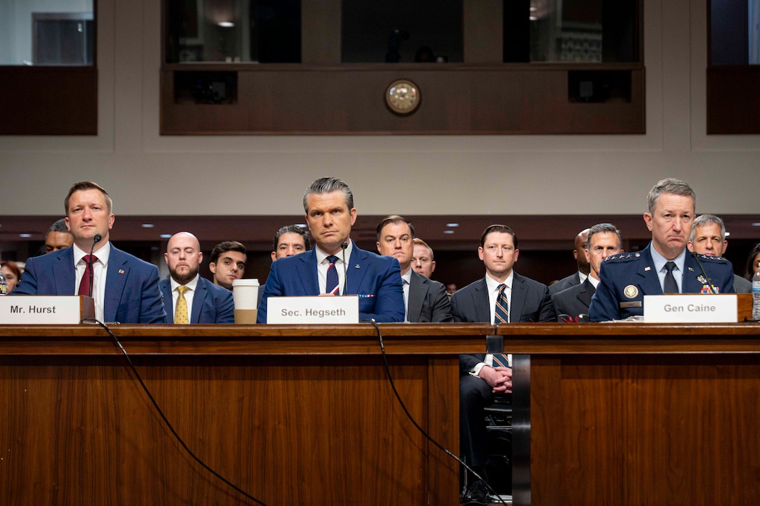 Two men in business suits and one man in a military uniform sit at a table in a Senate hearing room, with people sitting behind them.