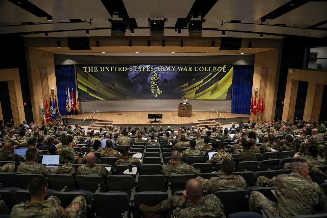 An auditorium filled with uniformed service members faces a stage where a speaker stands at a podium. Behind the podium is a large banner displaying the words 'THE UNITED STATES ARMY WAR COLLEGE' with an emblem at the center. Flags line both sides of the stage, and the audience occupies rows of tiered seating.