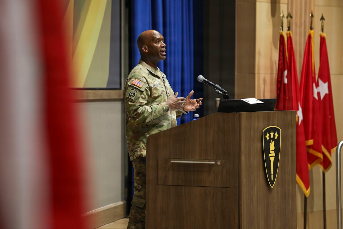 U.S. Army Gen. Xavier T. Brunson stands at a podium with a microphone, delivering remarks in an indoor auditorium. The podium displays an emblem with three stars and a torch. Several red military flags are positioned to the right, and a large curtain and wall paneling create the backdrop.