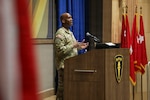 U.S. Army Gen. Xavier T. Brunson stands at a podium with a microphone, delivering remarks in an indoor auditorium. The podium displays an emblem with three stars and a torch. Several red military flags are positioned to the right, and a large curtain and wall paneling create the backdrop.