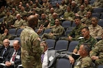 U.S. Army Gen. Xavier T. Brunson stands in front of an auditorium full of uniformed service members, appearing to address the audience while holding up three fingers. Rows of attendees sit in stadium-style seating, listening and taking notes.