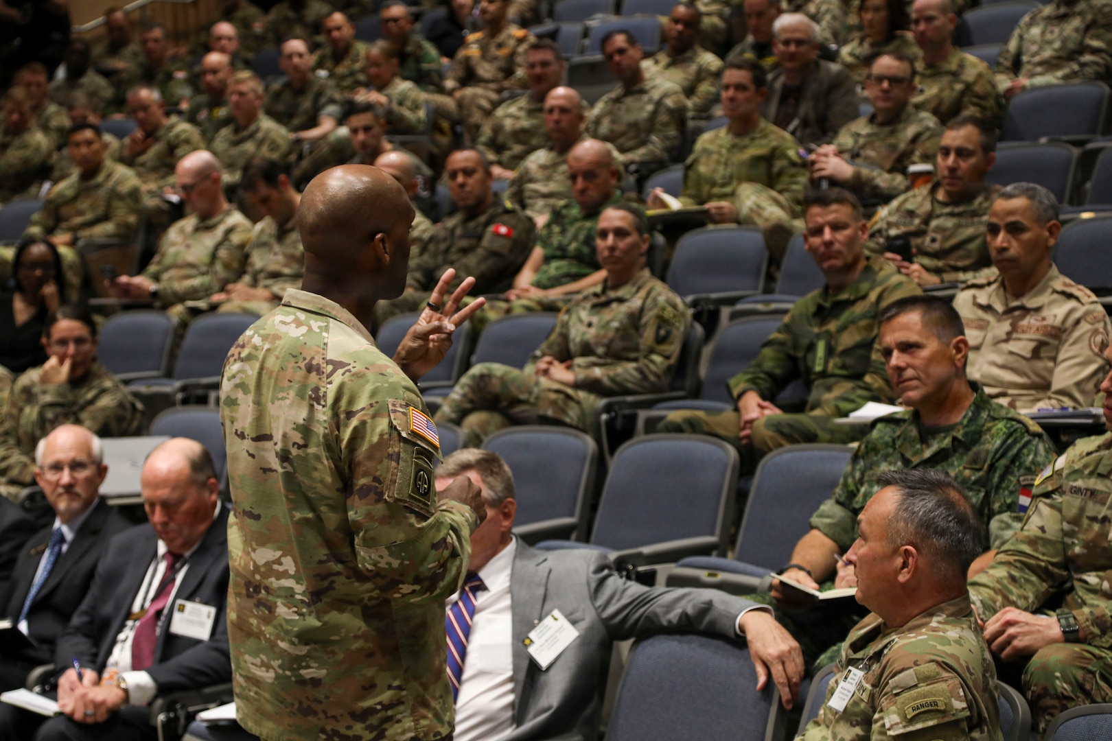 U.S. Army Gen. Xavier T. Brunson stands in front of an auditorium full of uniformed service members, appearing to address the audience while holding up three fingers. Rows of attendees sit in stadium-style seating, listening and taking notes.