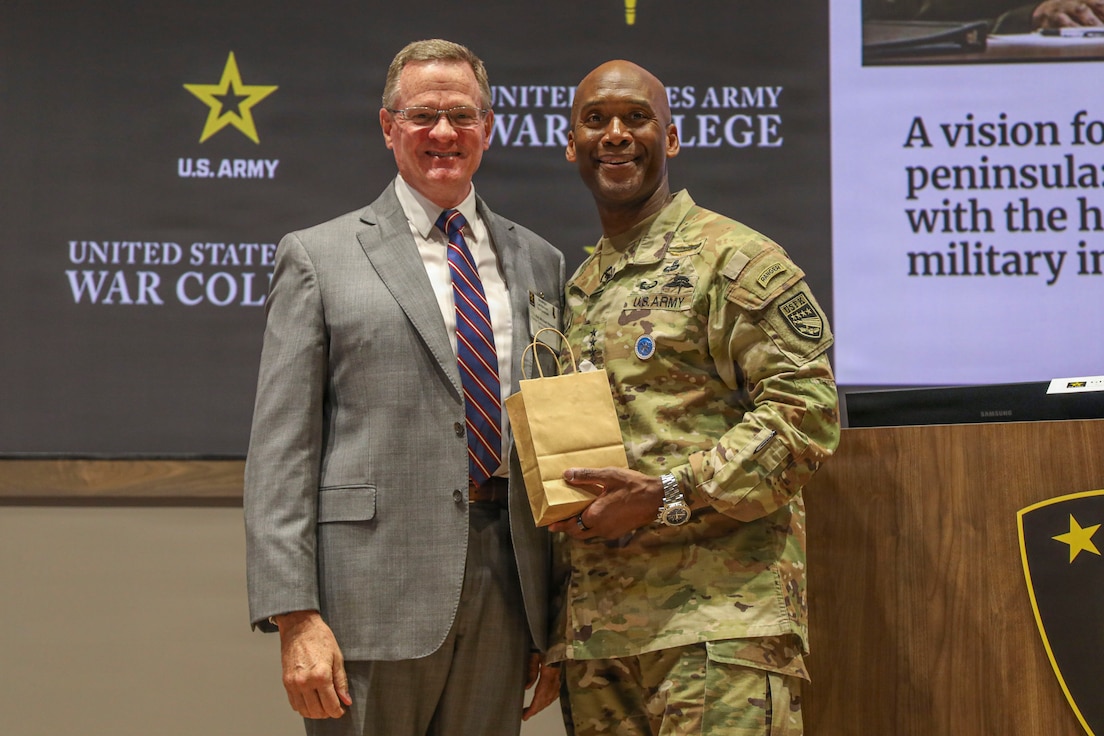 U.S. Army Gen. Xavier T. Brunson and U.S. Army Lt. Gen. (Ret.) Michael Howard stand side by side on a stage in front of a United States Army War College backdrop. Lt. Gen. (Ret.) Michael Howard is wearing a suit with a striped tie, and U.S. Army Gen. Xavier T. Brunson is wearing a U.S. Army uniform and holding a small brown gift bag.