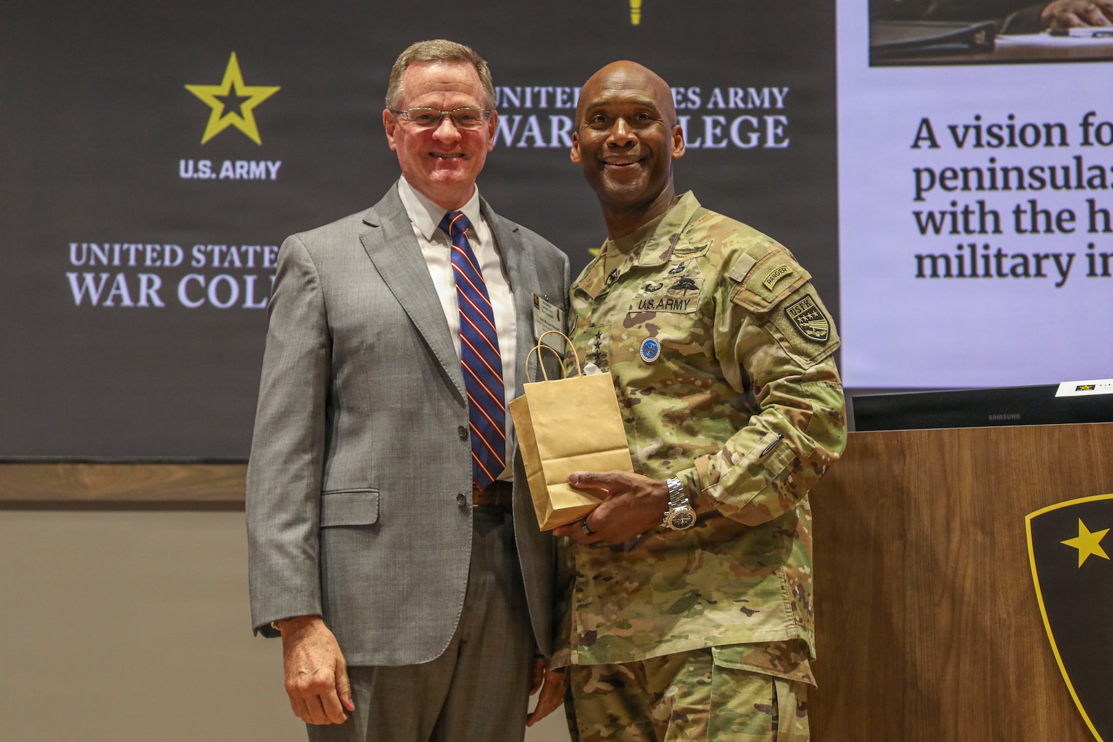 U.S. Army Gen. Xavier T. Brunson and U.S. Army Lt. Gen. (Ret.) Michael Howard stand side by side on a stage in front of a United States Army War College backdrop. Lt. Gen. (Ret.) Michael Howard is wearing a suit with a striped tie, and U.S. Army Gen. Xavier T. Brunson is wearing a U.S. Army uniform and holding a small brown gift bag.