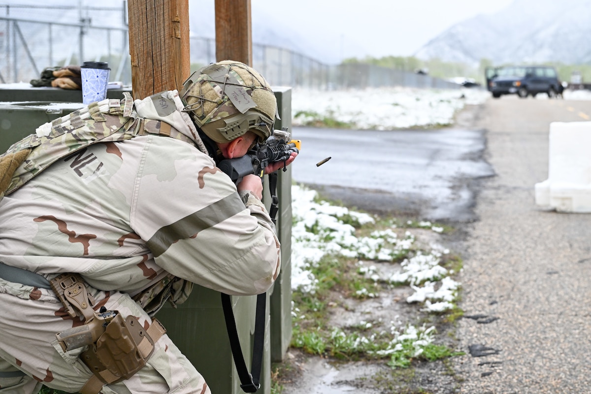 A uniformed airman aims and fires a rifle behind cover next to a road, with an ejected shell casing visible and snowy mountains in the background.