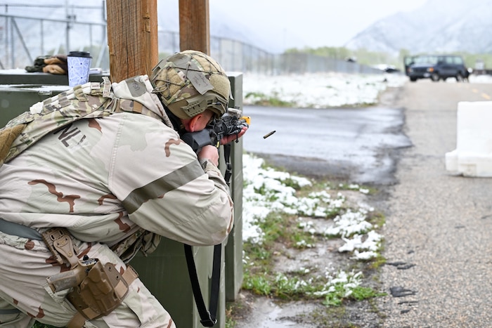 A uniformed airman aims and fires a rifle behind cover next to a road, with an ejected shell casing visible and snowy mountains in the background.