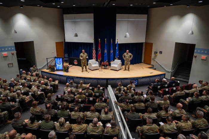 Two U.S. Airmen in OCP uniforms speak from a stage to an audience of other U.S. Airmen in flightsuits and OCP uniforms.
