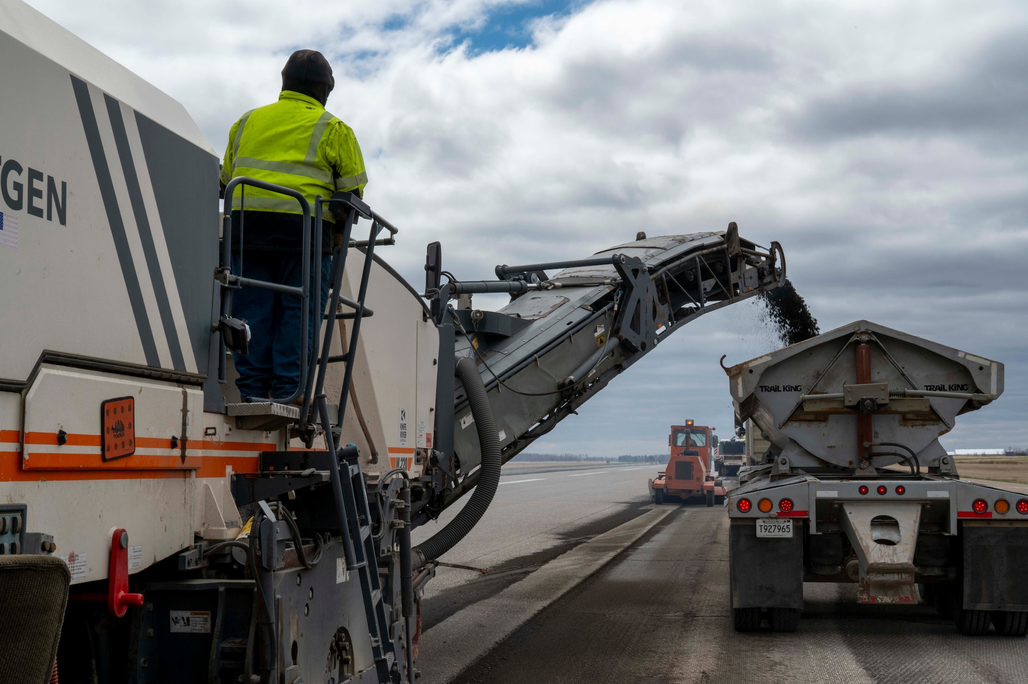A man in a green jacket oversees a construction truck pour asphalt into a dump truck.