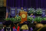 An individual in a military uniform stands at a wooden podium during a graduation ceremony, with purple banners behind the stage representing various university colleges such as the College of Arts and Sciences, the School of Nursing and Health Innovations, the School of Business, and the School of Engineering. Faculty members in academic regalia sit on the stage, and large floral arrangements surround the podium. Graduates in caps and gowns are seated in the foreground.
