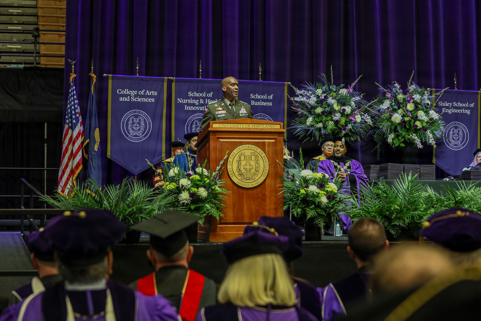 An individual in a military uniform stands at a wooden podium during a graduation ceremony, with purple banners behind the stage representing various university colleges such as the College of Arts and Sciences, the School of Nursing and Health Innovations, the School of Business, and the School of Engineering. Faculty members in academic regalia sit on the stage, and large floral arrangements surround the podium. Graduates in caps and gowns are seated in the foreground.