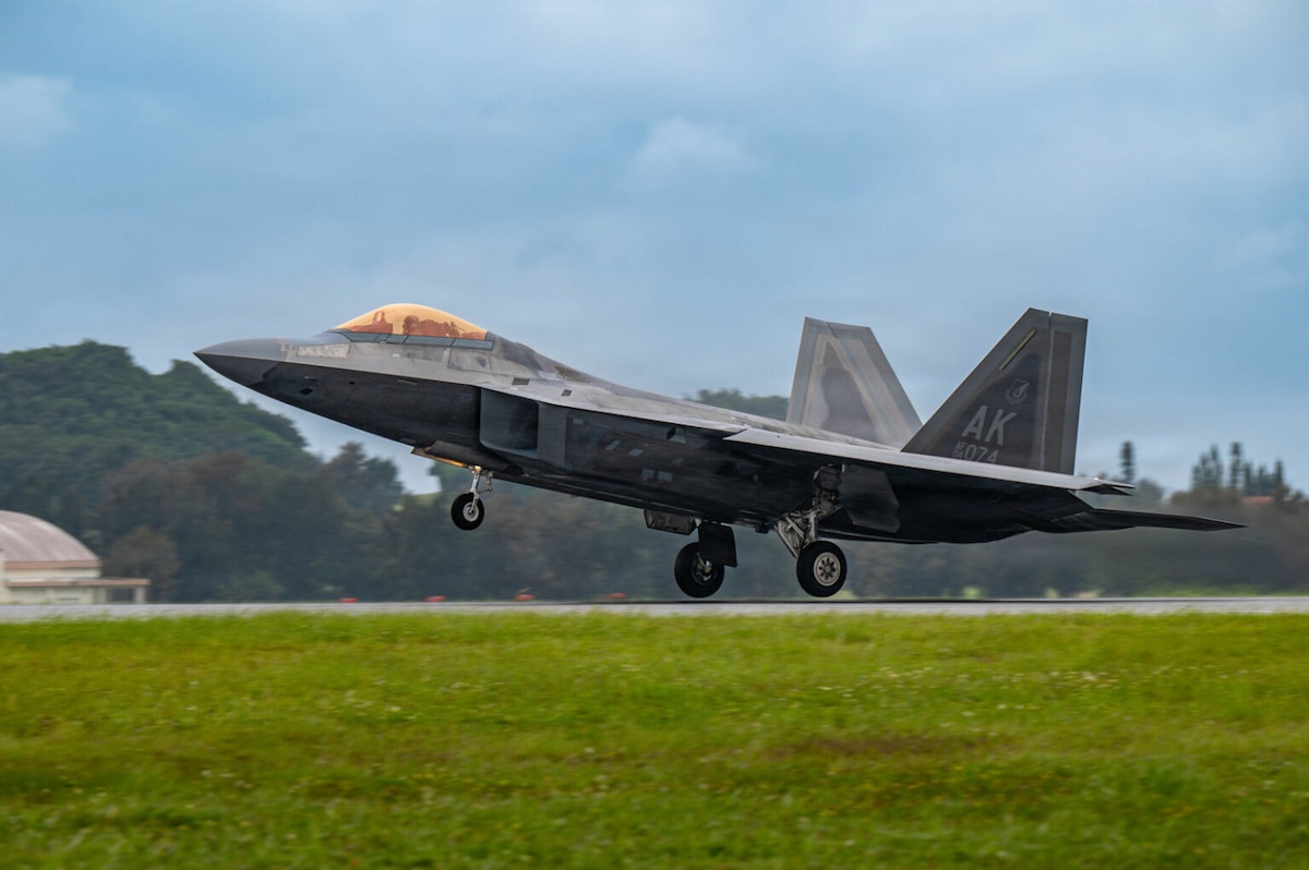 A military jet takes off from a runway, under a blue sky with trees in the background.