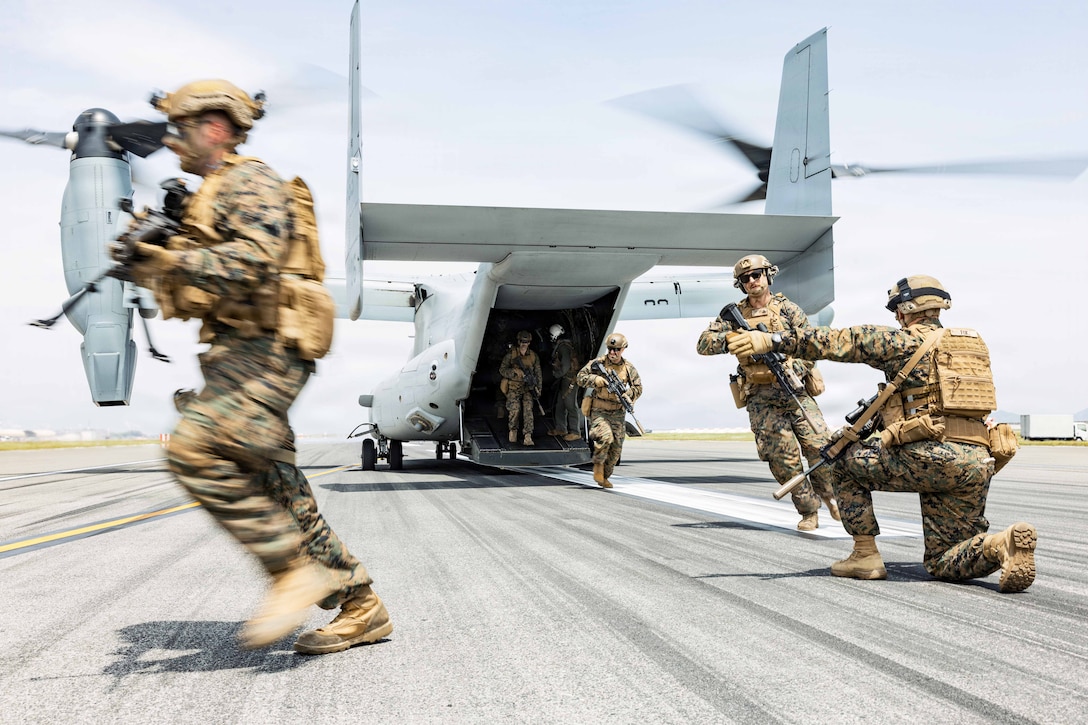 A Marine kneels to direct Marines in tactical gear as they run out of an aircraft on a tarmac during the day.