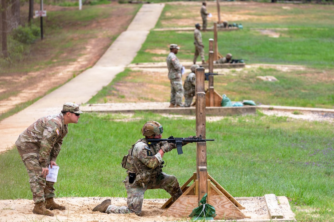 A soldier kneels in a sandbox to fire a weapon, leaning on a wooden structure as a fellow soldier observes.
