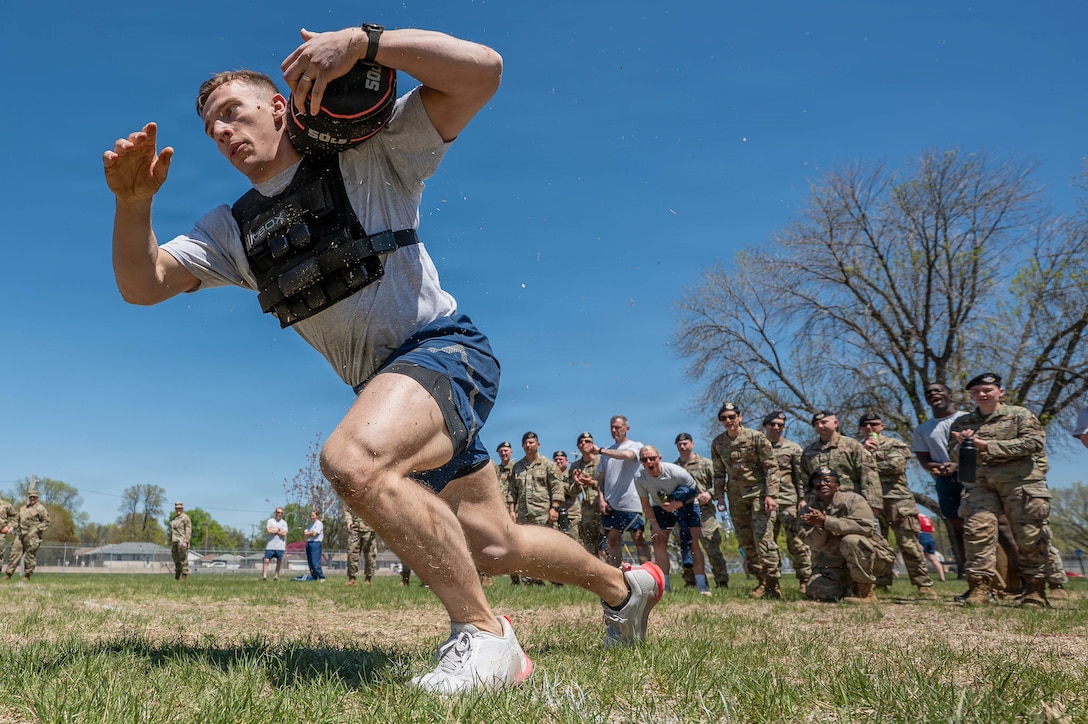 An airman wearing a weighted vest and carrying a weight over their shoulder prepares to run in a field against a blue sky as fellow service members cheer them on in the background.