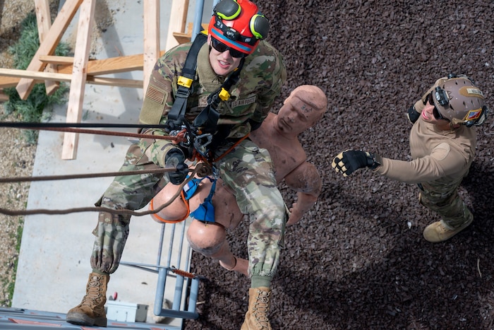 A man wearing a camouflage military uniform and helmet repels down the side of a building with a mannequin; there is another man in similar attire standing on the ground waiting for him.