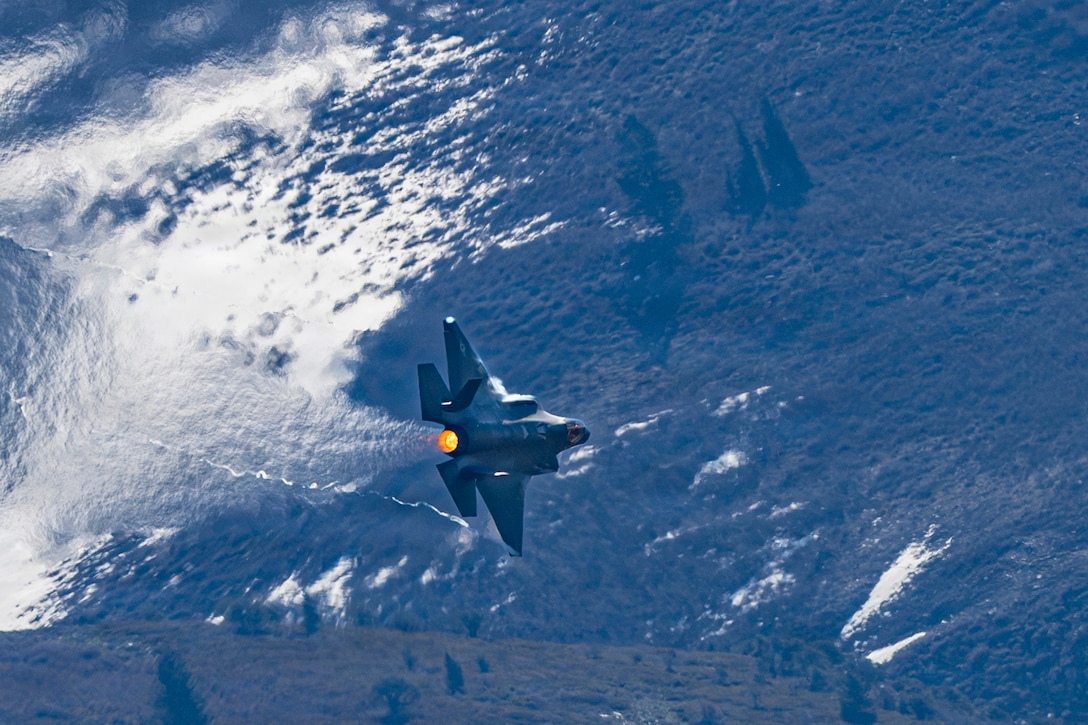 A fighter plane flies in front of a mountain that has patches of snow on it.