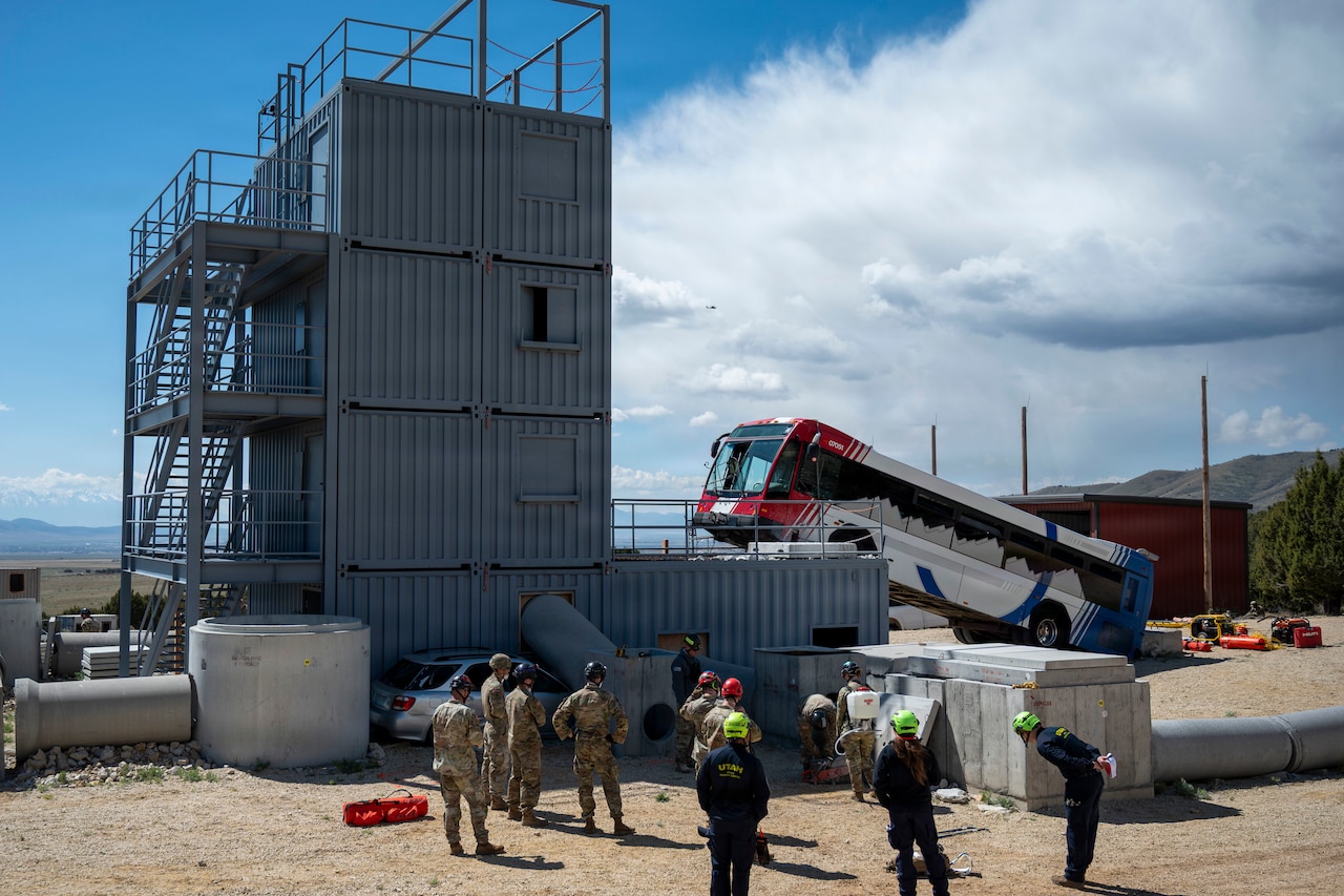 Eight people wearing camouflage military uniforms and helmets, and four people in coveralls and helmets, stand outside near a tall building as they prepare for training; there is a bus in the background with its front wheels on the side of the building.