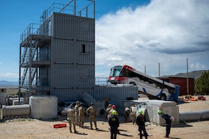Eight people wearing camouflage military uniforms and helmets, and four people in coveralls and helmets, stand outside near a tall building as they prepare for training; there is a bus in the background with its front wheels on the side of the building.