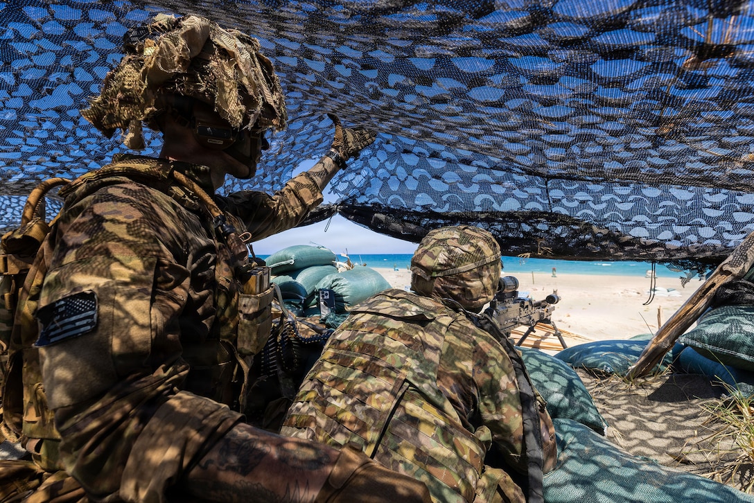 Two soldiers under a tarp on a beach shoot targets, with one soldier holding a weapon and the other assisting.