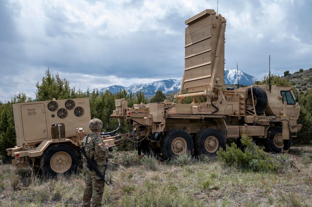 A man wearing a camouflage military uniform stands outside in a wooded area looking at a large military radar.