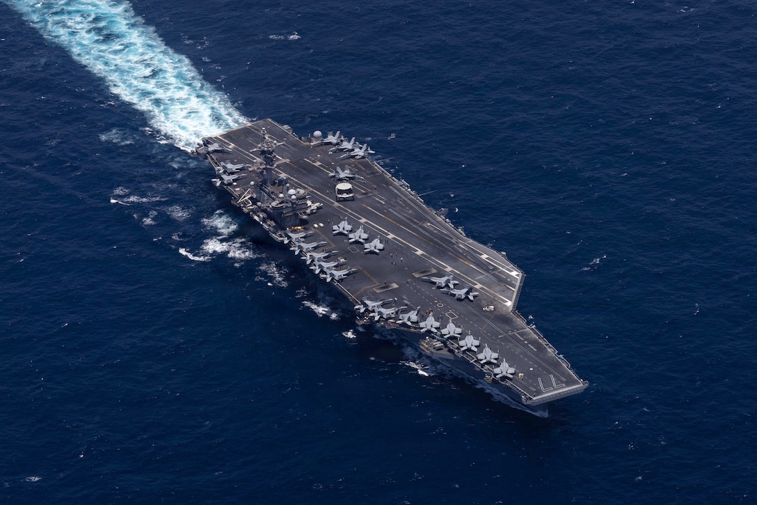 Fighter planes sit on an aircraft carrier sailing in deep blue water, viewed from above.