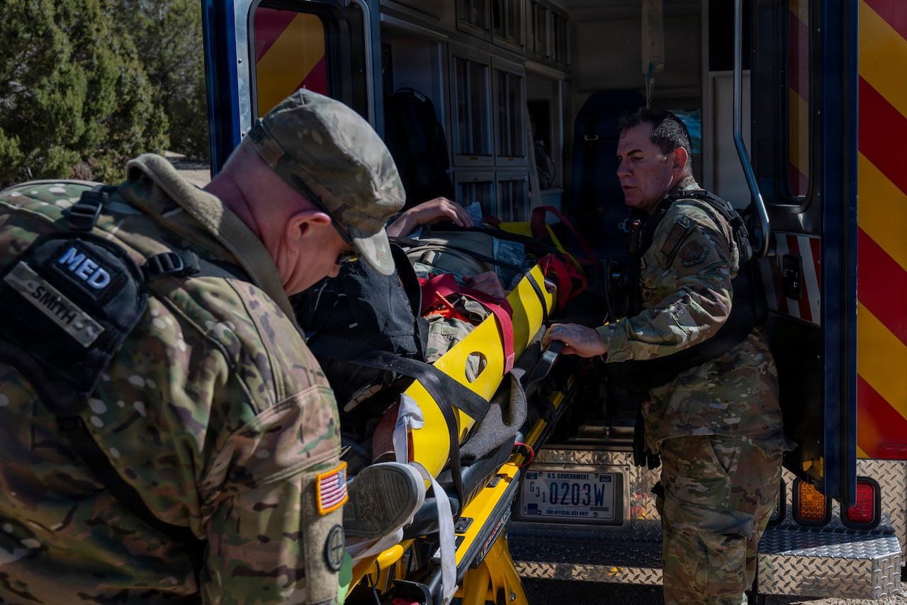 Two men wearing camouflage military uniforms load a man on a stretcher into an ambulance.