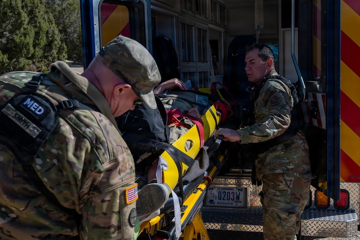 Two men wearing camouflage military uniforms load a man on a stretcher into an ambulance.