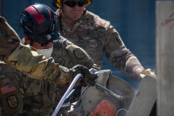 A man wearing a camouflage military uniform and helmet uses a saw to cut a slab of concrete while two men in similar attire assist him.
