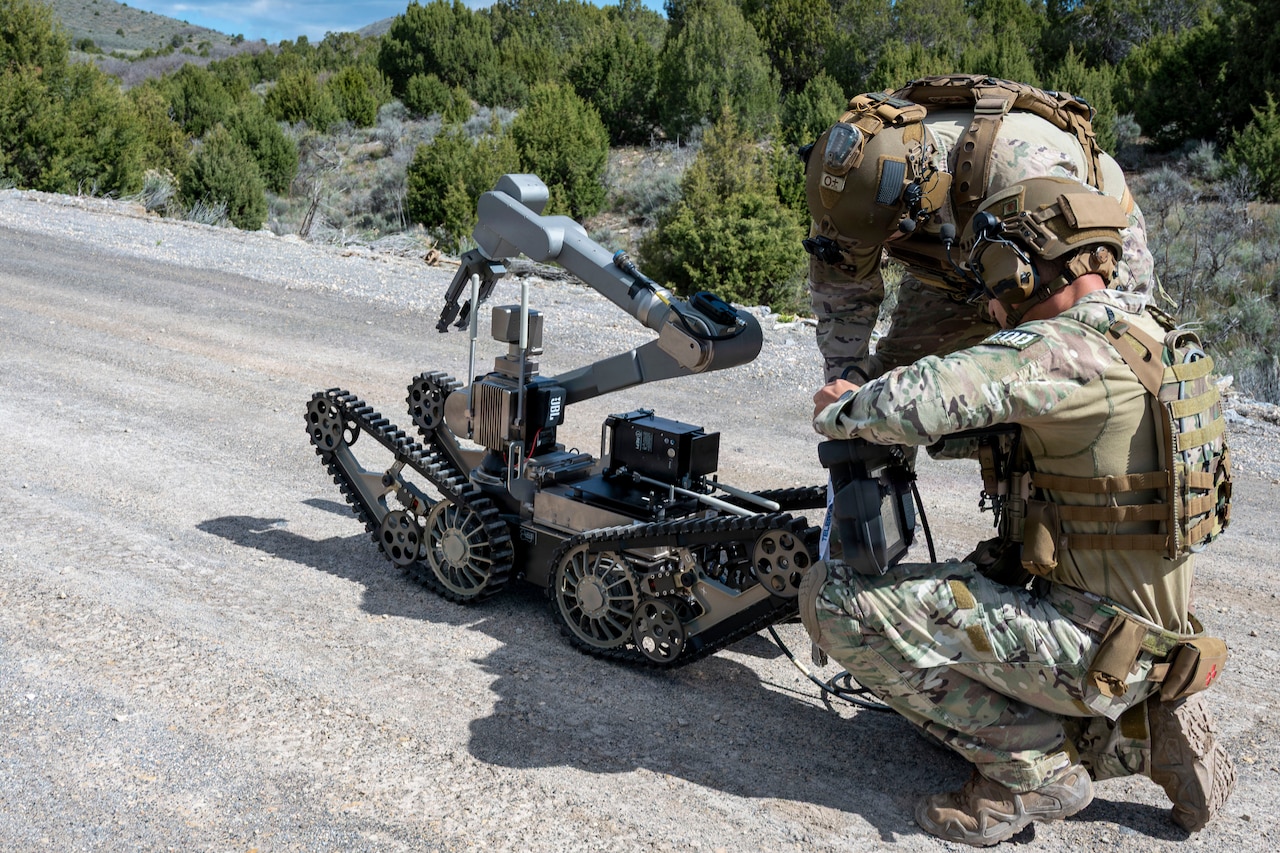 Two men wearing camouflage military uniforms and tactical gear work on a bomb robot outside on a gravel road; there are trees and mountains in the background.